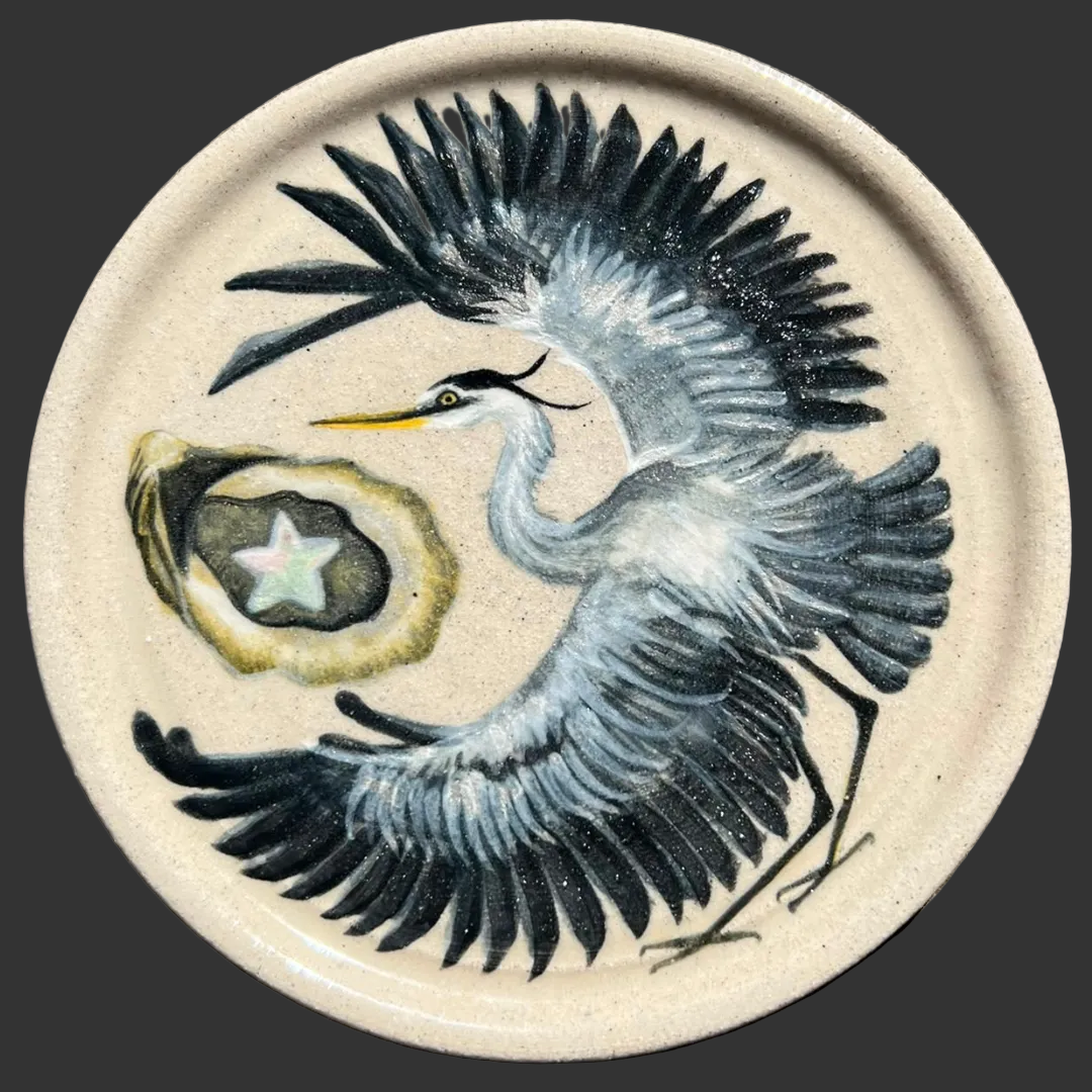A round ceramic plate featuring a painted heron with outstretched dark blue-black wings and a grey-and-white body, alongside an oyster with a star pearl.
